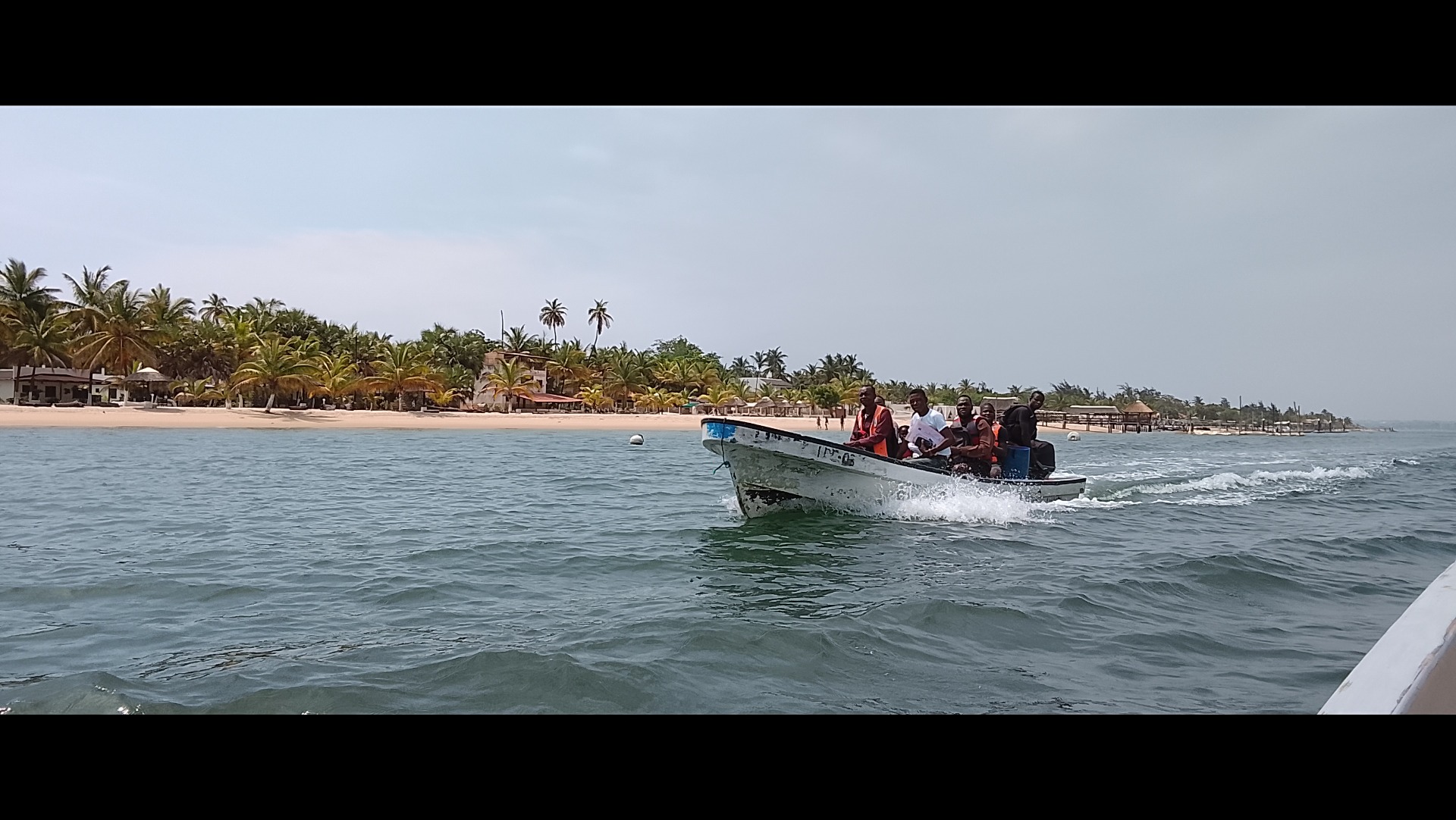 Small boat with several people speeds across ocean toward a palm-lined beach under cloudy sky.