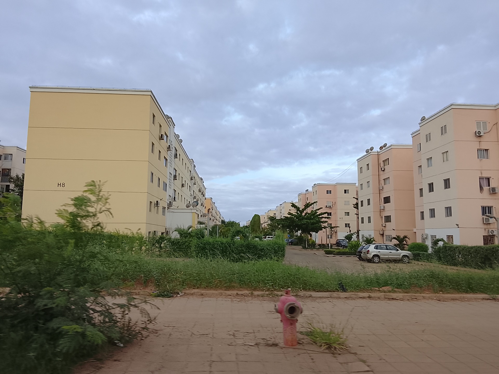 Prédios residenciais bege e brancos, carros estacionados e vegetação, sob céu nublado na Centralidade Kilamba em Luanda
