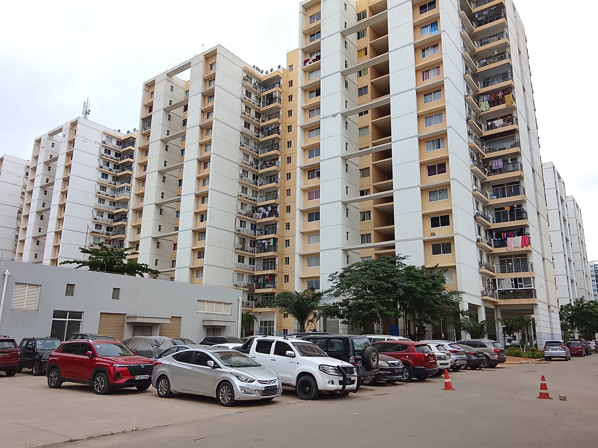 Prédio residencial alto na centralidade do Zango em Luanda com varandas, janelas e roupas, carros estacionados em frente e árvores no pátio.