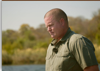 Bald man in a green shirt stands outdoors, looking down with a thoughtful expression.
