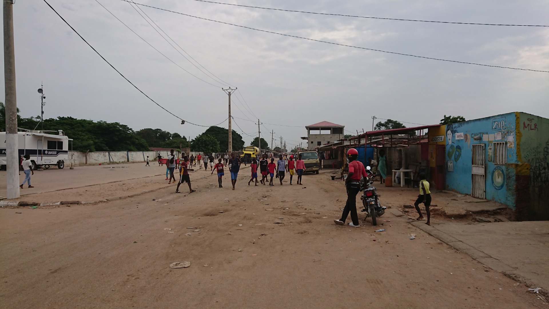 pais: Angola. Pátio de rua com crianças correndo, veículos estacionados e fios elétricos sob céu nublado.