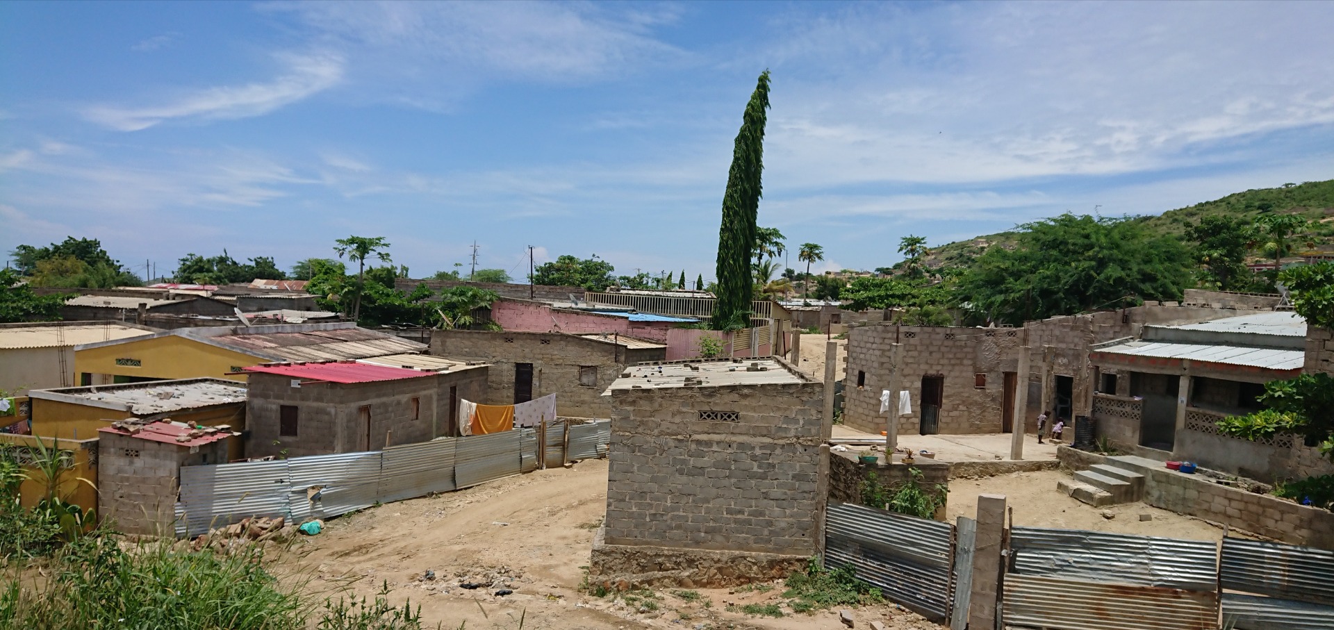 Pais: Angola. Vista de um bairro com casas de alvenaria, cercas metálicas e uma árvore alta sob céu azul.