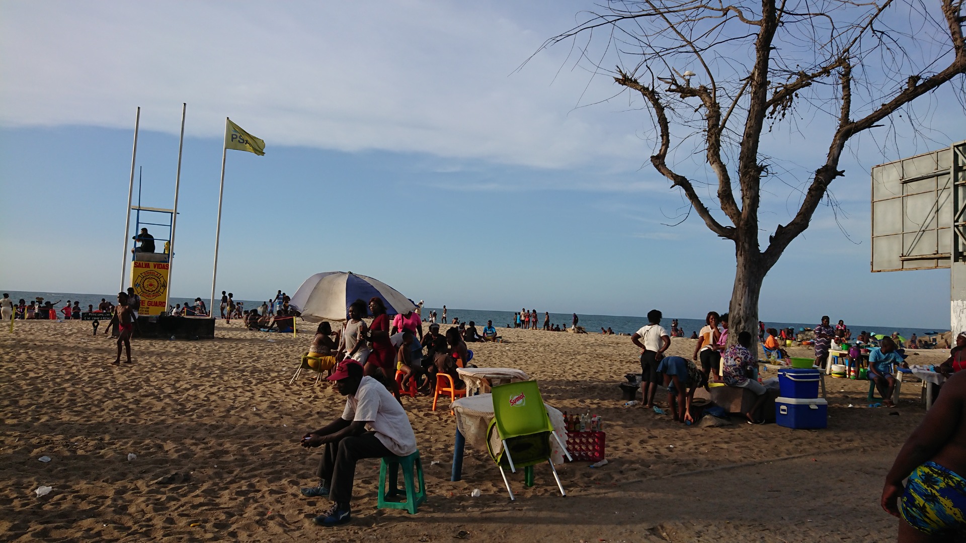 Angola. Praia movimentada com pessoas sentadas, barraca branca e árvore ao lado, céu nublado e mar ao fundo.