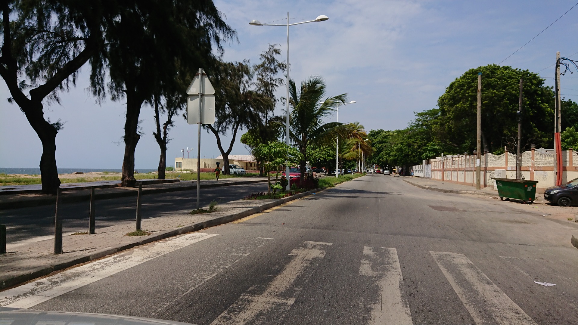 Angola. Rua costeira arborizada com palmeiras, postes de iluminação e carros estacionados sob céu parcialmente nublado.