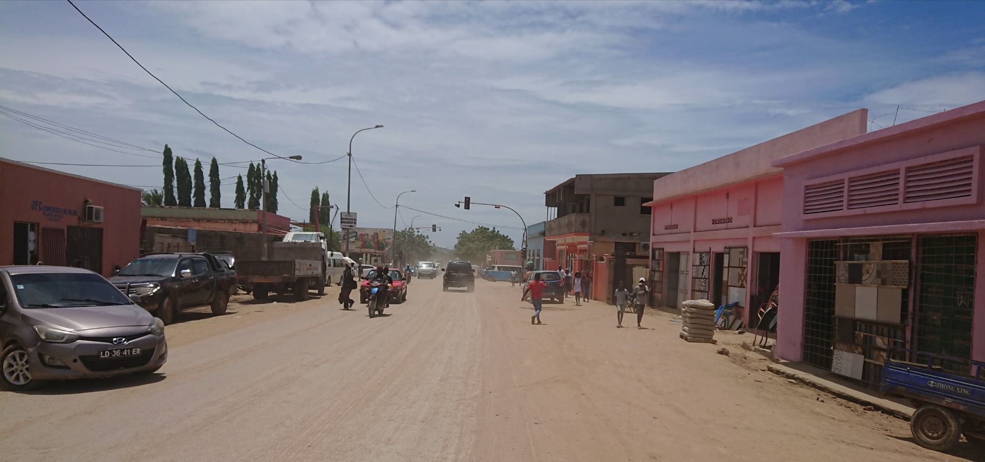Angola - Rua movimentada com carros estacionados, motocicletas e pedestres entre prédios industriais sob céu nublado.