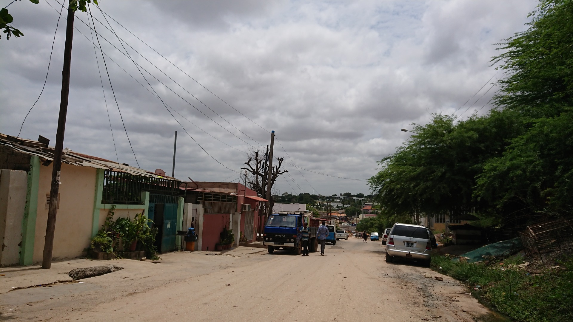 Angola - Rua residencial com carros estacionados, postes e árvores verdes sob céu nublado.
