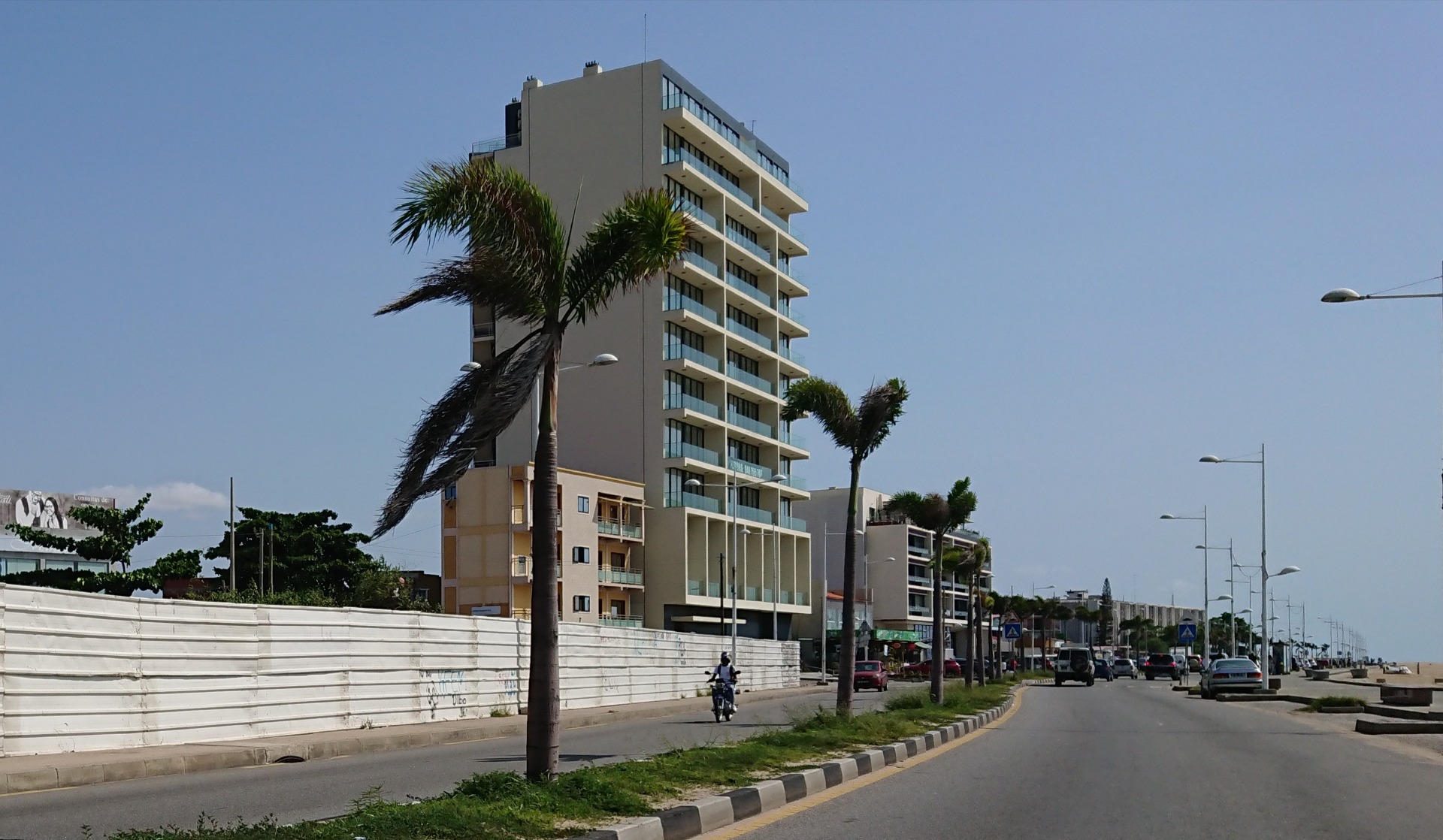 Luanda- Angola - Prédio alto residencial moderno com varandas, palmeiras e avenida com carros ao fundo, céu azul.