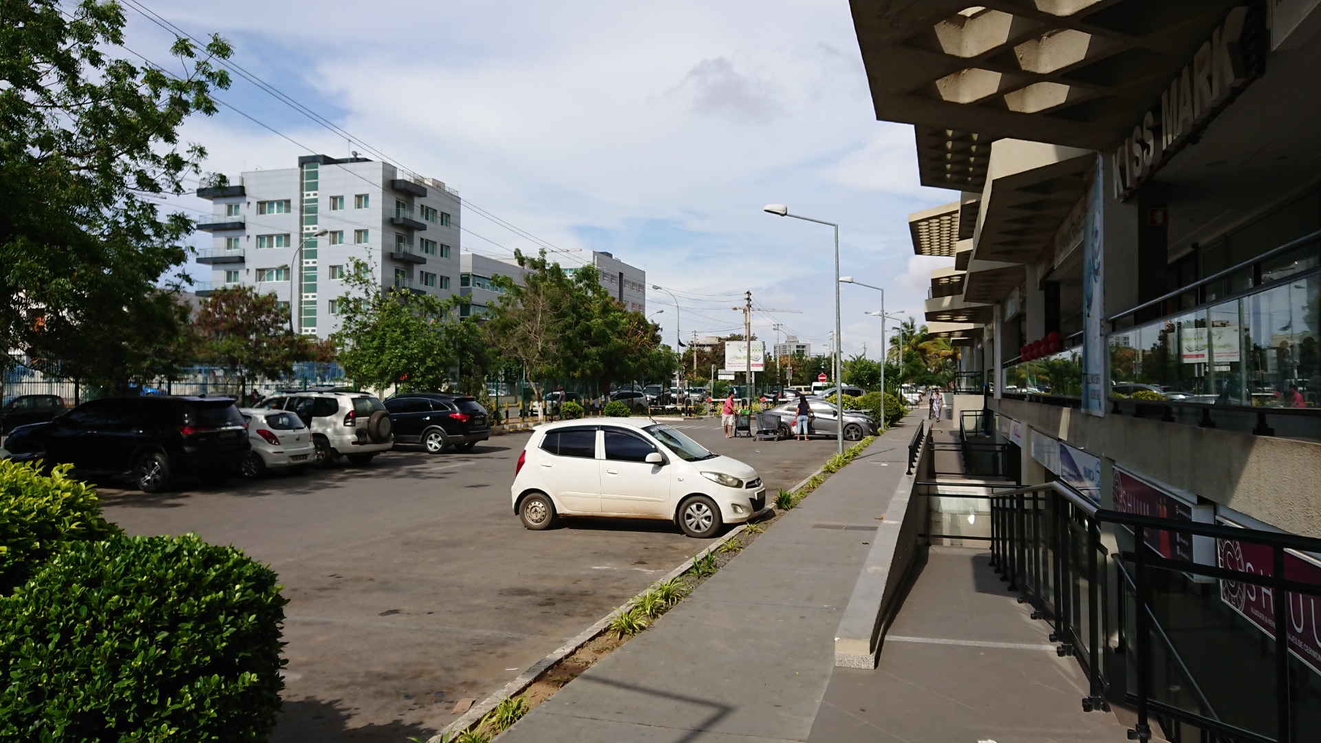 Angola - Luanda - Estacionamento ao lado de prédio moderno, carros alinhados, árvores e calçada sob marquise com luzes.