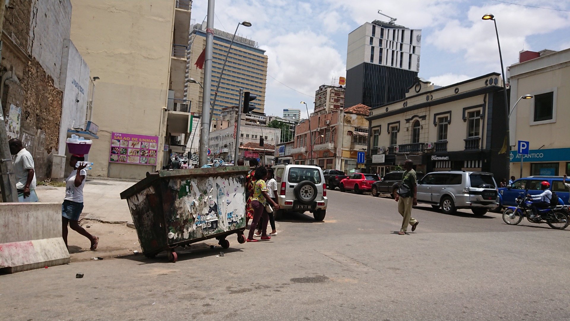 Luanda - Angola - Rua movimentada com carros e pedestres, prédios altos ao fundo e bandeiras ao céu parcialmente nublado.