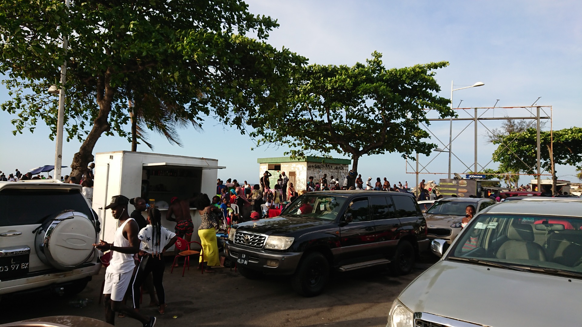 Angola - Pessoas reunidas em estacionamento ao ar livre, carros alinhados e árvores grandes sombreando o local.