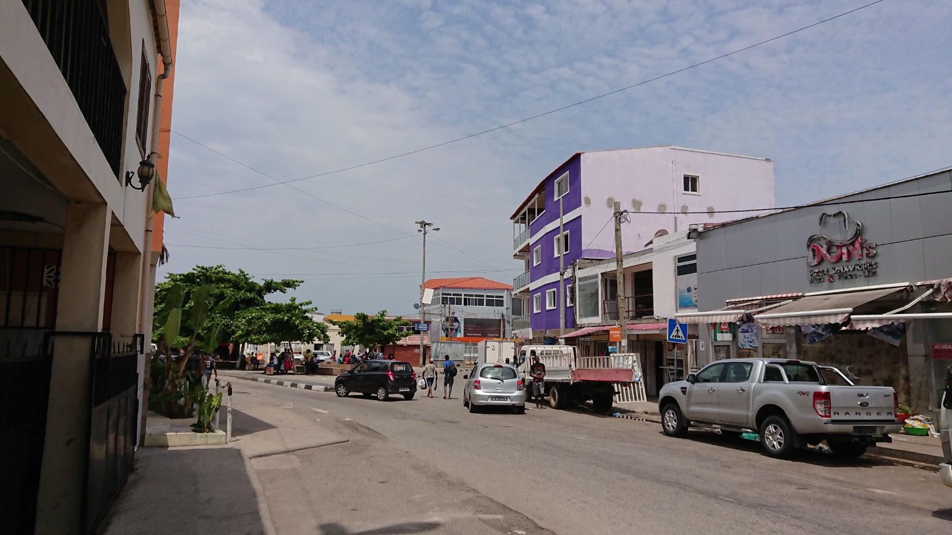 Angola - Rua movimentada com carros estacionados, prédio roxo e branco, lojas e céu parcialmente nublado.