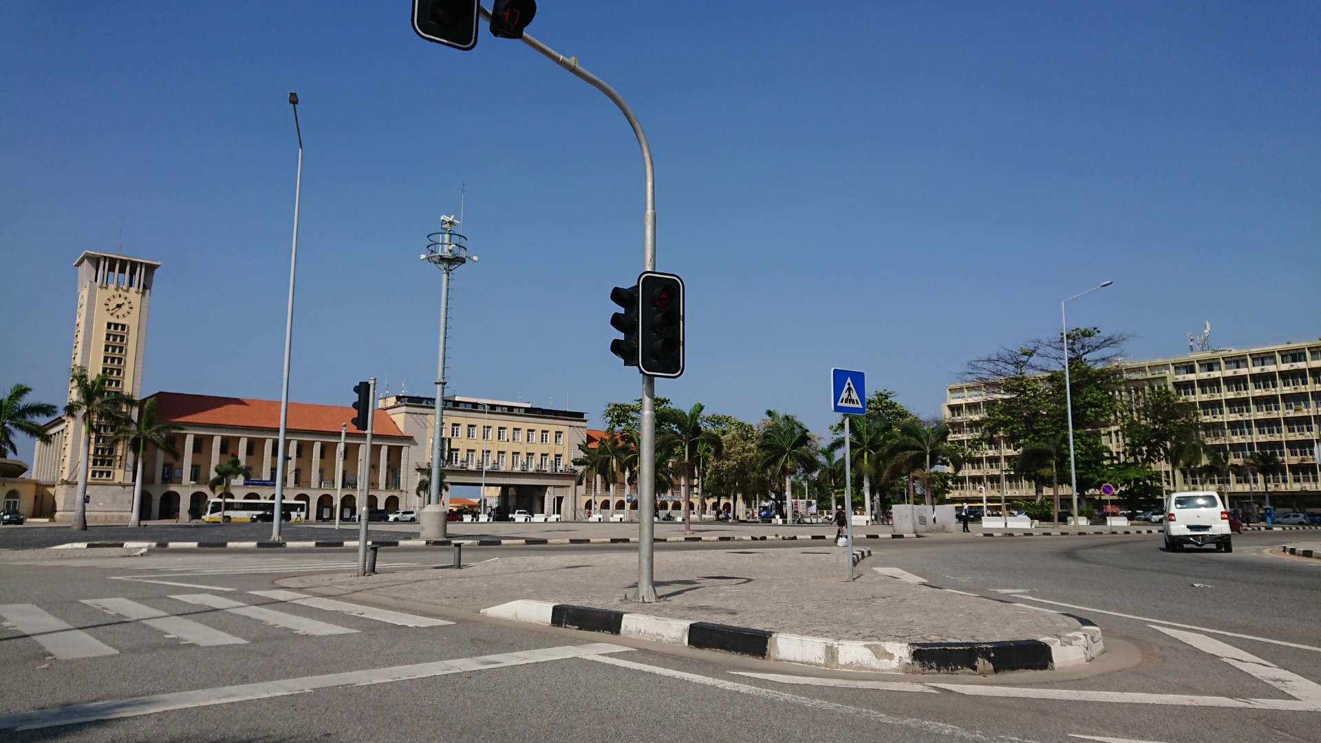 Port of Luanda- Urban intersection with traffic lights, crosswalk sign, palm trees, and colonial buildings under a clear blue sky.