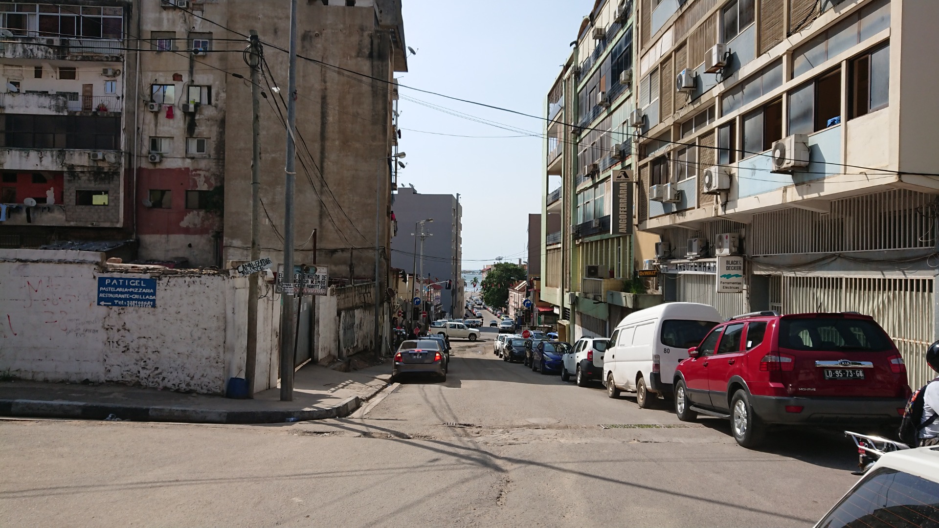 Urban street in Luanda with parked cars, tall apartment buildings, overhead wires, and distant traffic under clear sky.