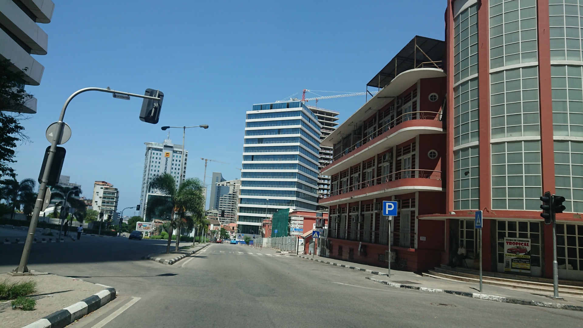 Angola - Luanda - Urban street with tall office buildings, construction cranes, and a red building beside a parking sign.