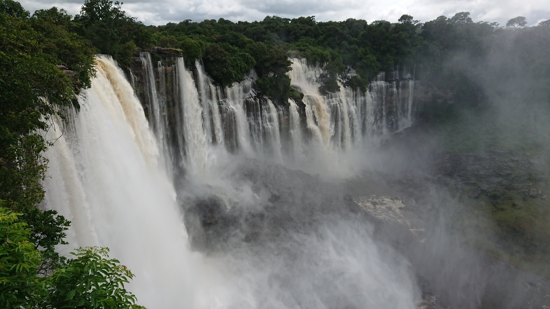 Massive waterfall cascades over rocky cliffs into mist, surrounded by dense green forest under cloudy Kalandula Falls sky.